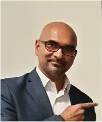 A man with glasses and a beard, wearing a suit and white shirt, points to the right.